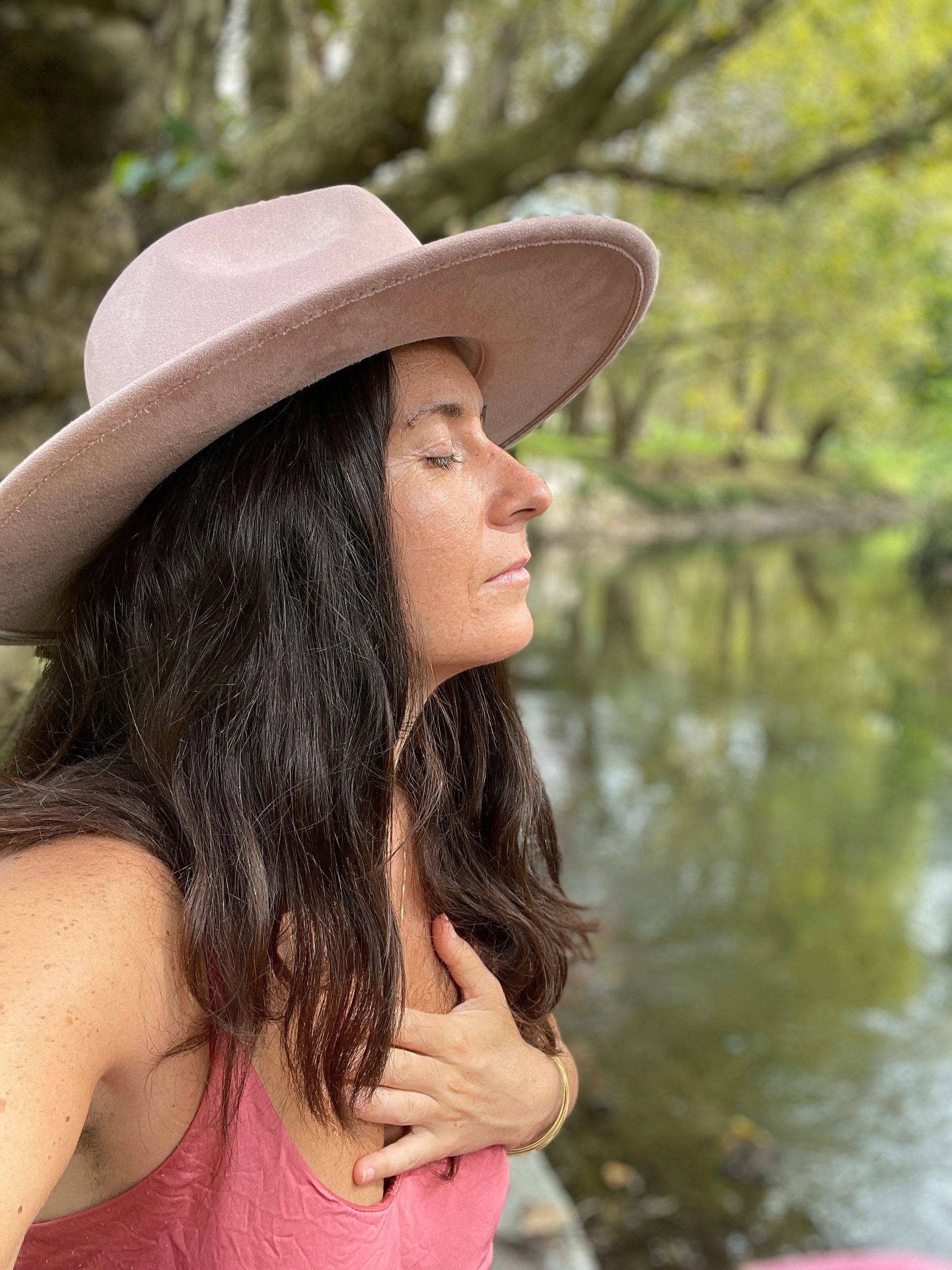 Woman in a pink hat meditates peacefully by a river.