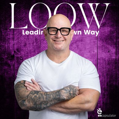 Smiling bald man with tattoos and glasses stands confidently against a purple wooden background.