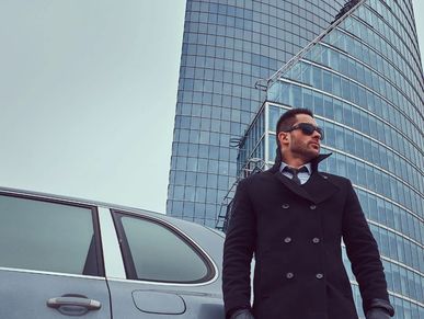 Man in sunglasses and coat standing by a car in front of a modern glass building.