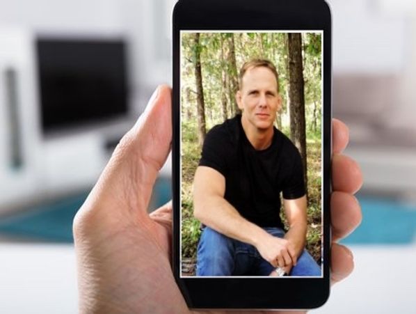 A man in a black shirt sitting outdoors, shown on a phone screen.