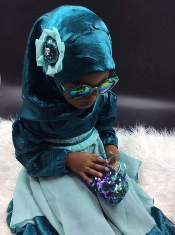 A young girl in a teal dress and hijab holding a sequined purse.