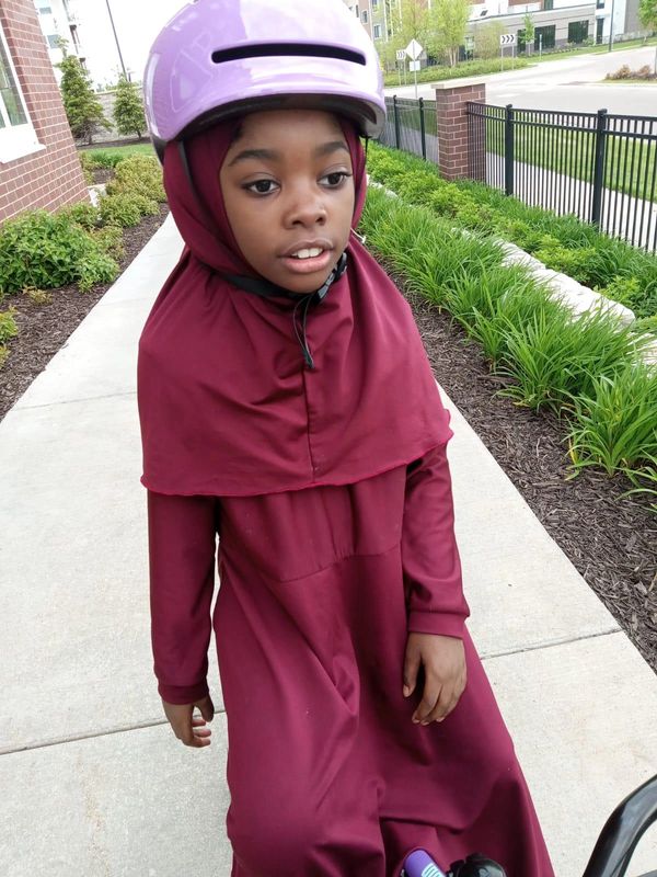 Young girl wearing a maroon dress and hijab with a purple helmet outdoors.