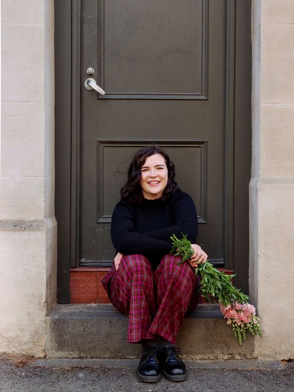 Sam is sitting on a step of a doorway smiling at the camera. She is holding a bouquet of snapdragons