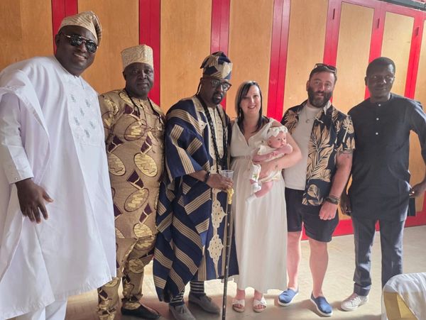 Group of six adults and a baby posing indoors, some in traditional African attire.