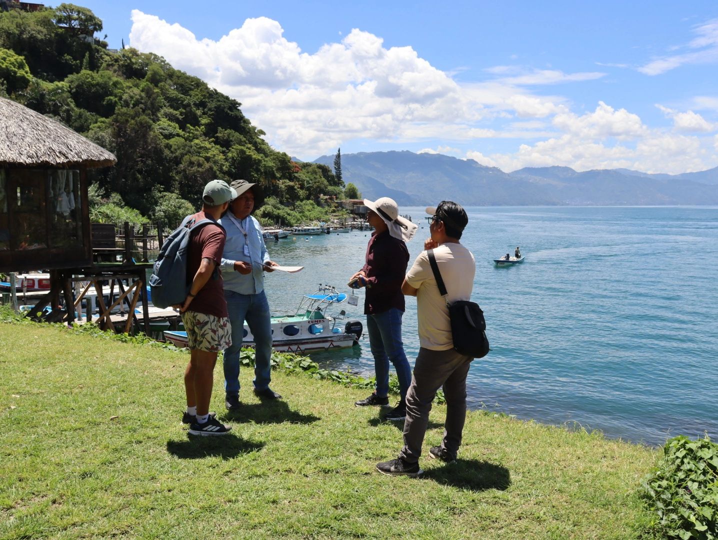 Cuatro personas conversan junto a un lago con montañas al fondo.