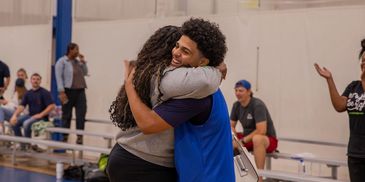 Two people warmly hugging in a gymnasium setting.
