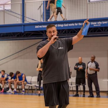 Man speaking into a microphone at a basketball game in a gym.