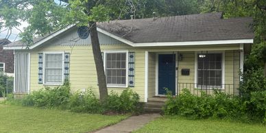 Small yellow house with blue door and windows, surrounded by greenery.