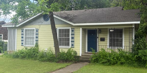 Small yellow house with blue door and windows, surrounded by greenery.