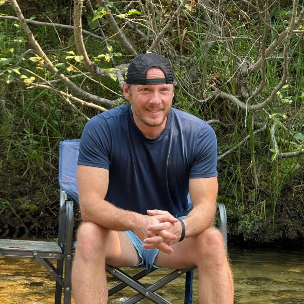 Man sitting on a chair near a natural water body with greenery.
