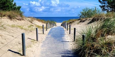 A sandy path leads to a beach under a blue sky with clouds.