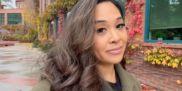 Woman with long curly hair posing outdoors against a building with autumn foliage.