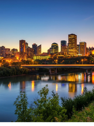 Edmonton skyline at dusk with buildings lit, a bridge over the river, and trees in the foreground.