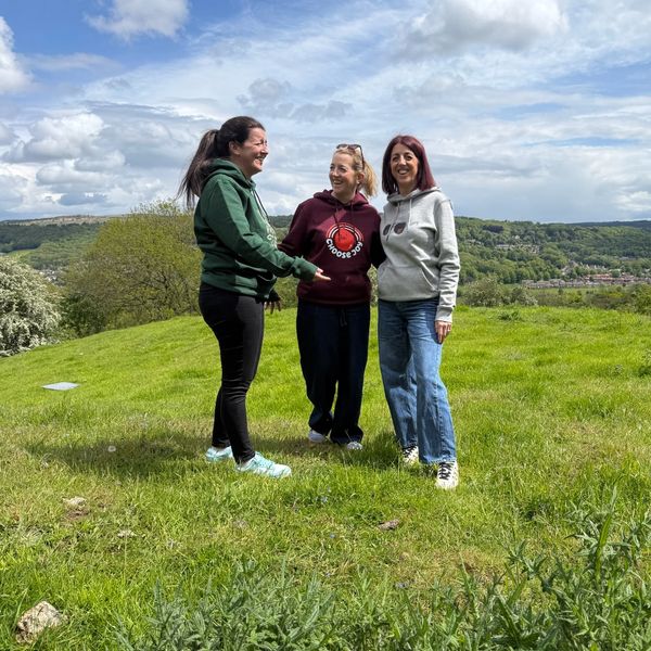 Three women smiling and chatting on a grassy hill under a partly cloudy sky.