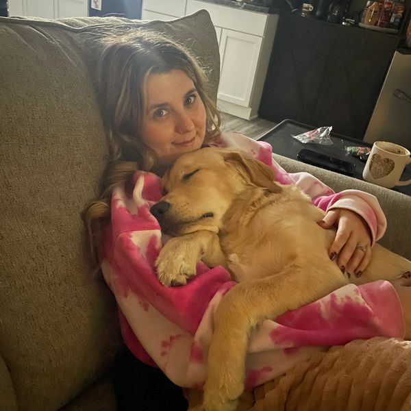 A woman in a pink tie-dye hoodie cuddles a sleeping golden retriever on a couch.