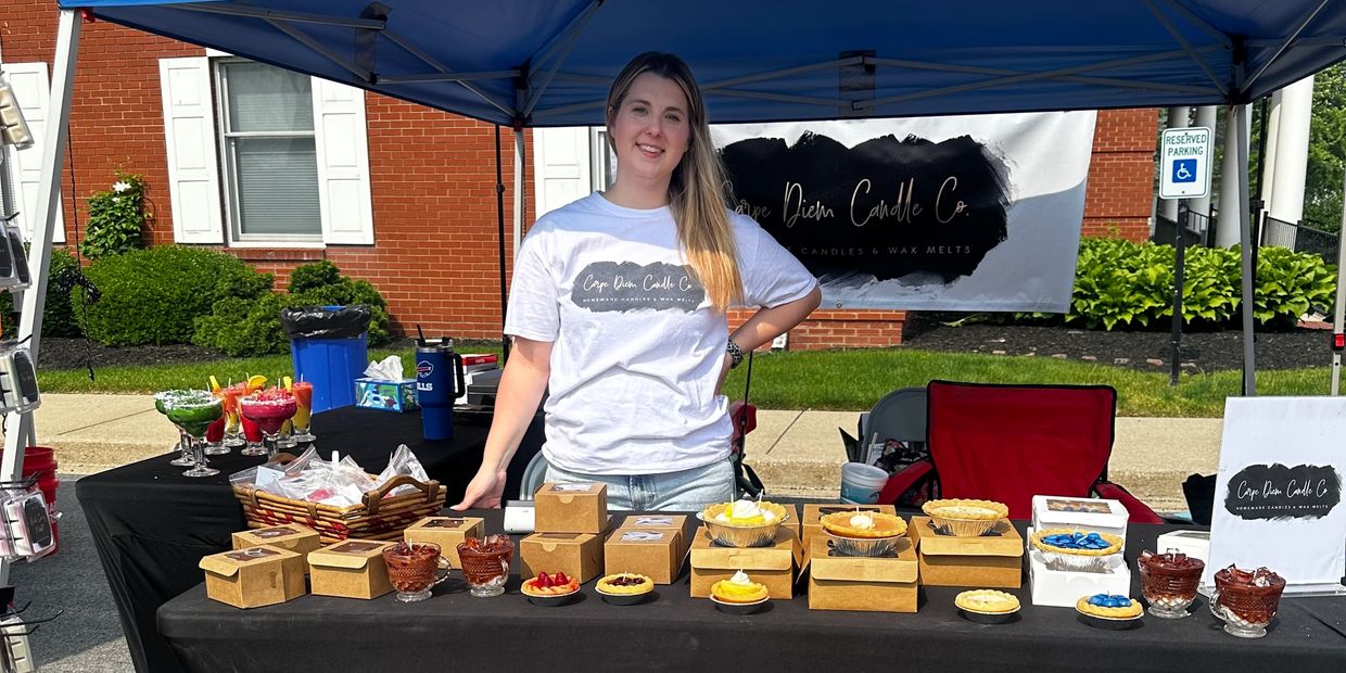 Woman selling handmade candles and wax melts at an outdoor market stall.