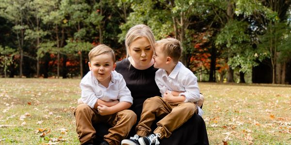 A woman with two young boys sitting outdoors, one boy kissing her cheek.