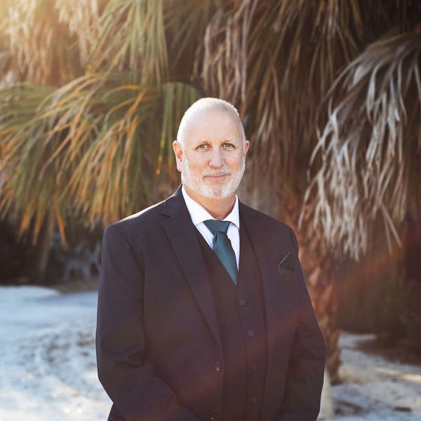 A man in a dark suit standing outdoors with palm trees in the background.