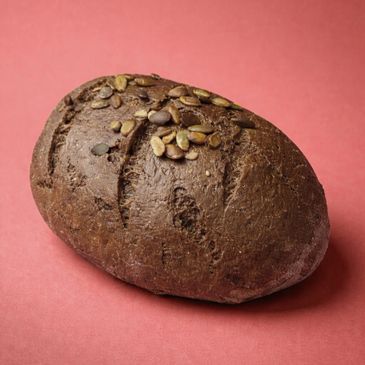A loaf of dark bread topped with pumpkin seeds on a pink background.