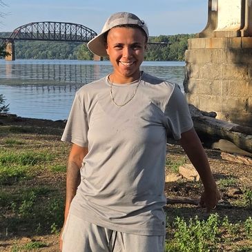 Person smiling by a riverside with a bridge in the background.