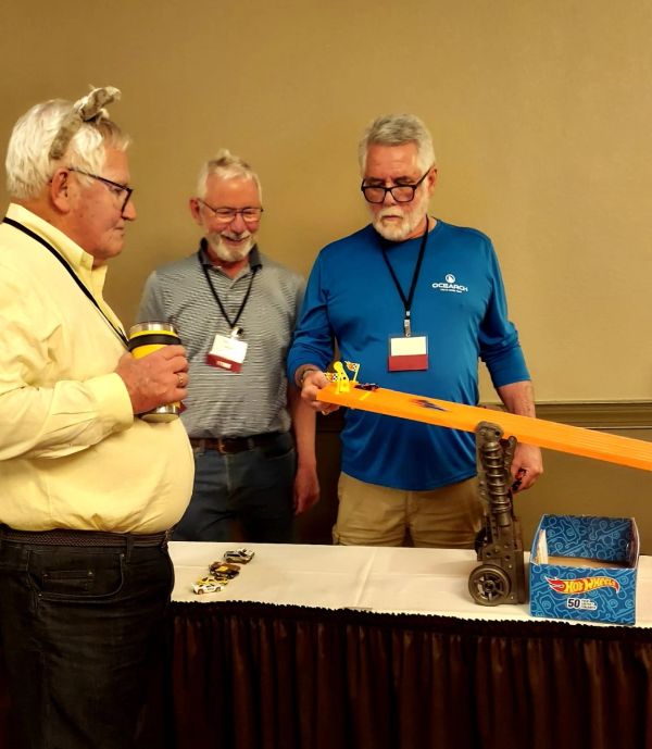 Three men engaged in a Hot Wheels car race on an orange track indoors.