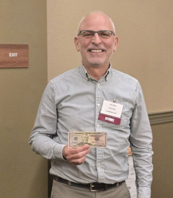 Smiling man with glasses holding a twenty-dollar bill, wearing a retiree badge.