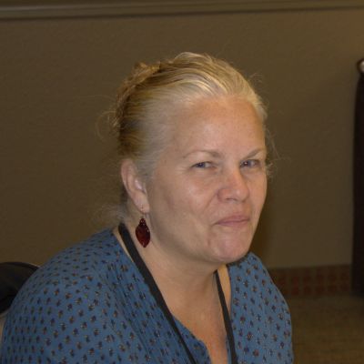 Smiling woman with blonde hair wearing a blue patterned top and maroon earrings.