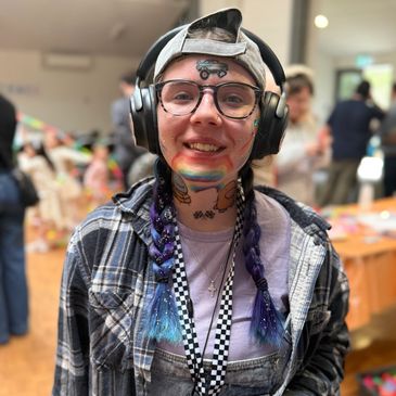 Person with face and neck face paint, headphones, glasses, and braided hair smiles indoors.