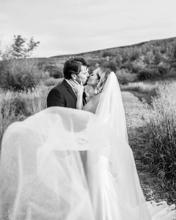 A bride and groom share a kiss outdoors with her veil flowing dramatically.