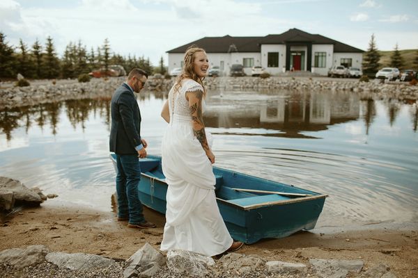 Bride and groom by a lake with a blue boat and a large house in the background.