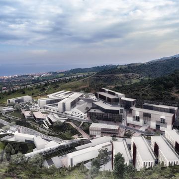 A large modern campus nestled in green hills near a coastal town under a partly cloudy sky.