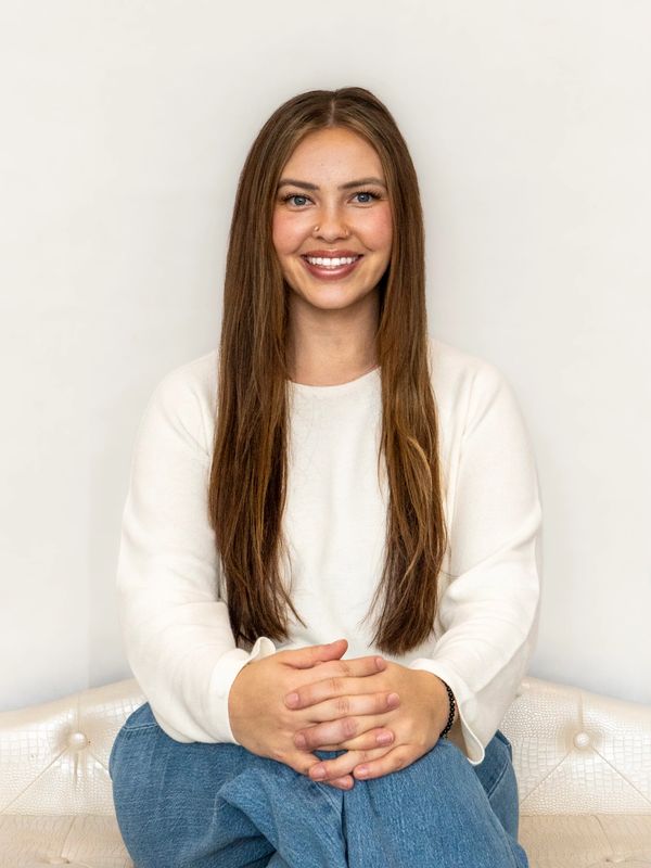 Young woman with long brown hair smiling, sitting with hands clasped.