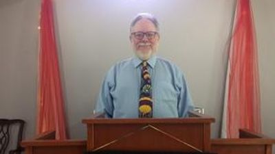 A man in a blue shirt and colorful tie standing behind a wooden podium.