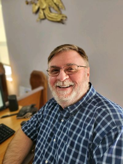 Smiling man in a blue checkered shirt sitting at a desk.