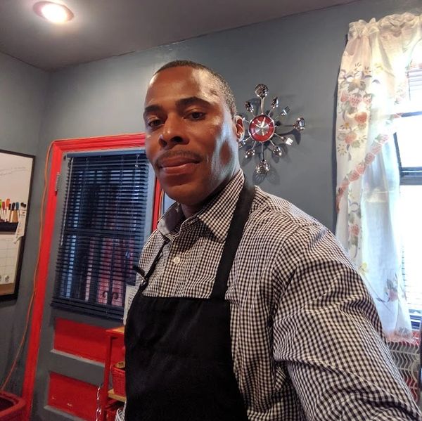 Man in checkered shirt and black apron in a cozy kitchen.