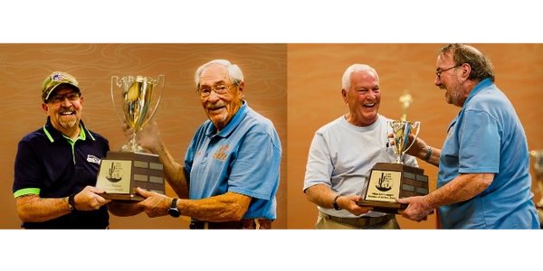Two pairs of elderly men smiling while holding trophies together against an orange backdrop.