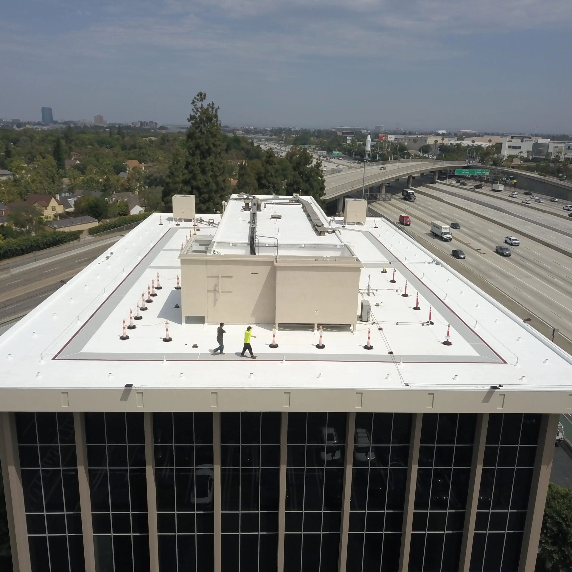 Two workers walk on a white rooftop of a tall building near a busy highway.