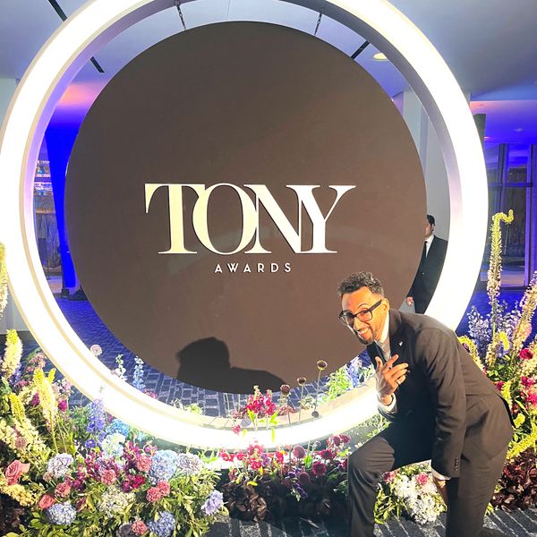 Man posing happily beside a lit Tony Awards sign surrounded by flowers.