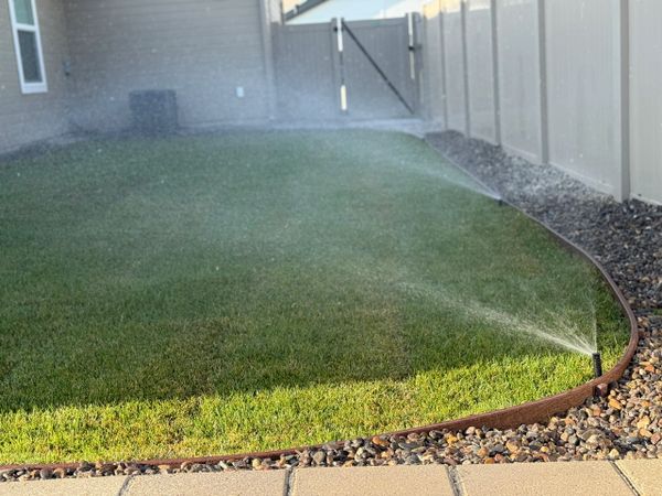 A sprinkler watering a green lawn bordered by stones and a fence.