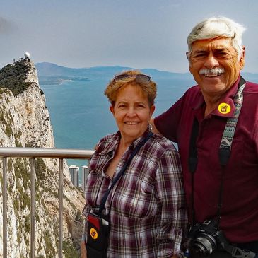 Elderly couple smiling with a scenic cliff and ocean backdrop.