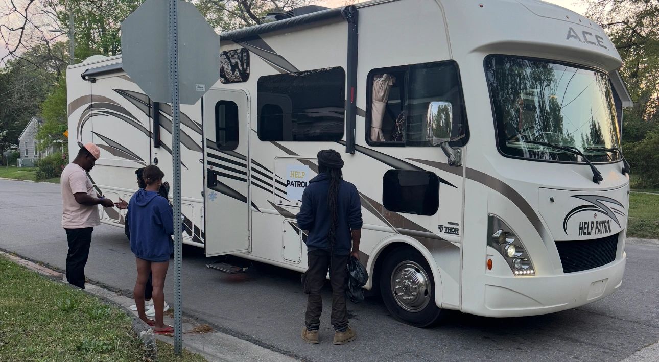 People standing near a white Help Patrol RV on a suburban street.