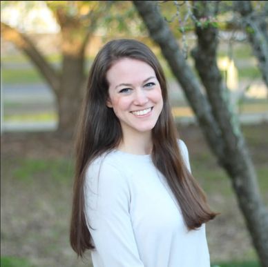 Young woman with long brown hair smiling outdoors by trees.