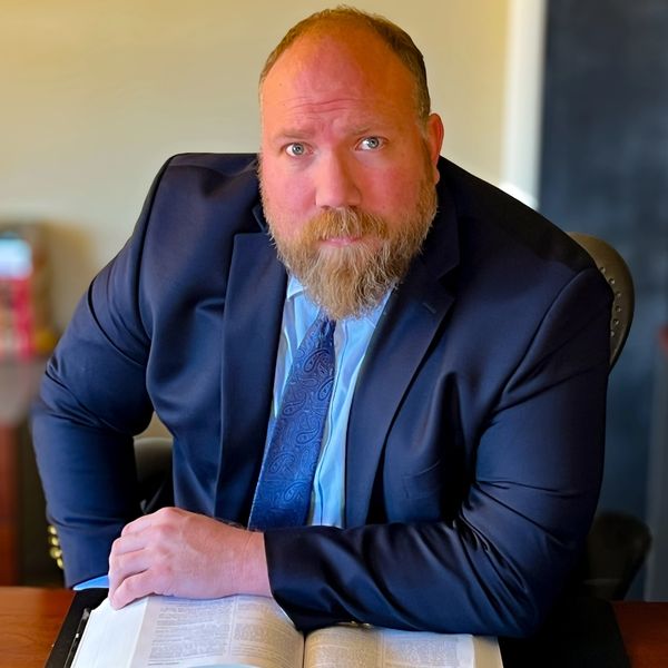 Man in a suit reading a book at a desk, looking thoughtfully at the camera.