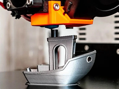 A 3D printer creating a small boat model in gray filament.