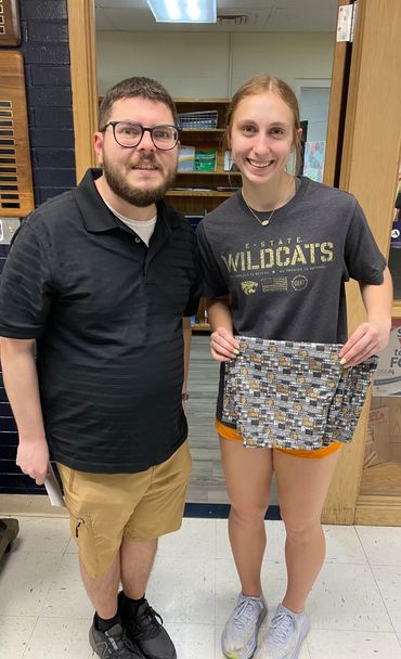 Two people smiling, one holding patterned fabric, standing indoors.