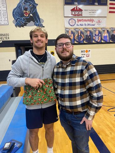 Two men posing together in a gymnasium, one holding a colorful item.