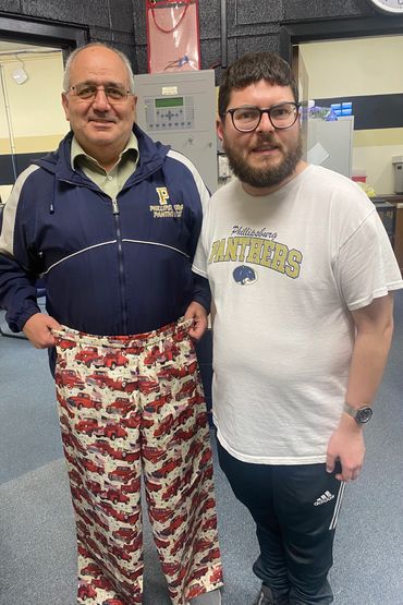 Two men standing indoors, one holding patterned pants with red trucks.