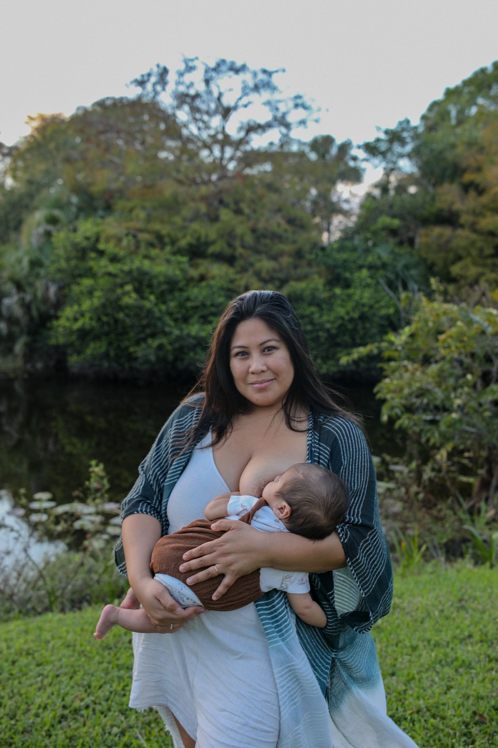 A woman breastfeeding her baby outdoors near greenery.