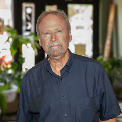 Older man in a dark shirt standing indoors with plants.