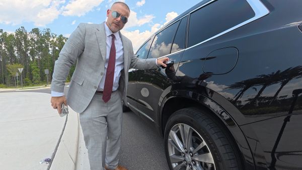 Man in a gray suit opening a black car door on a sunny day.